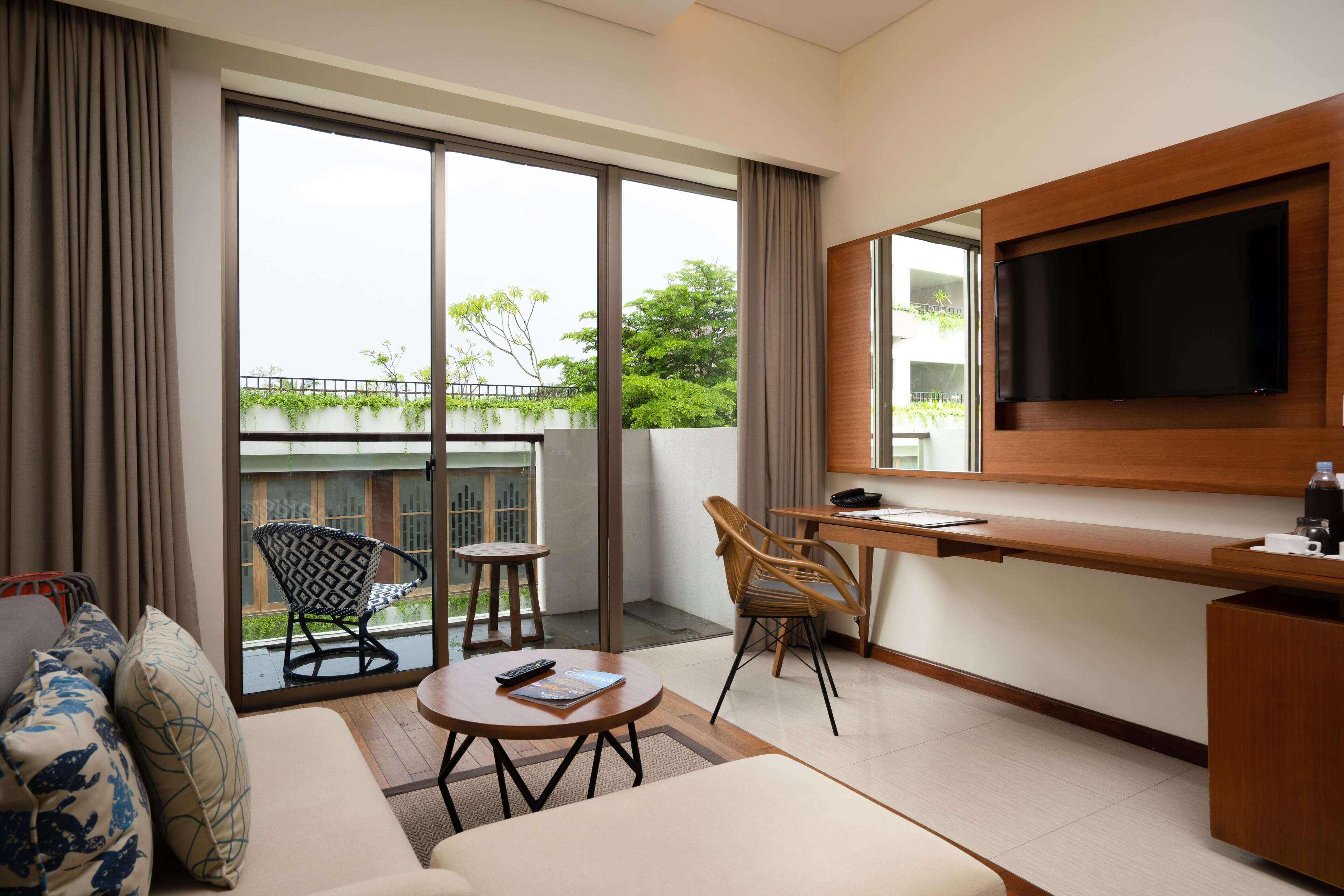 Habitación de hotel moderna con un amplio ventanal corredero que da a un balcón, un televisor de pantalla plana en la pared, un sofá con cojines estampados, una pequeña mesa redonda y dos sillas.