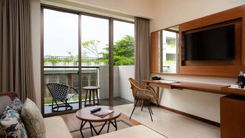 Habitación de hotel moderna con un amplio ventanal corredero que da a un balcón, un televisor de pantalla plana en la pared, un sofá con cojines estampados, una pequeña mesa redonda y dos sillas.