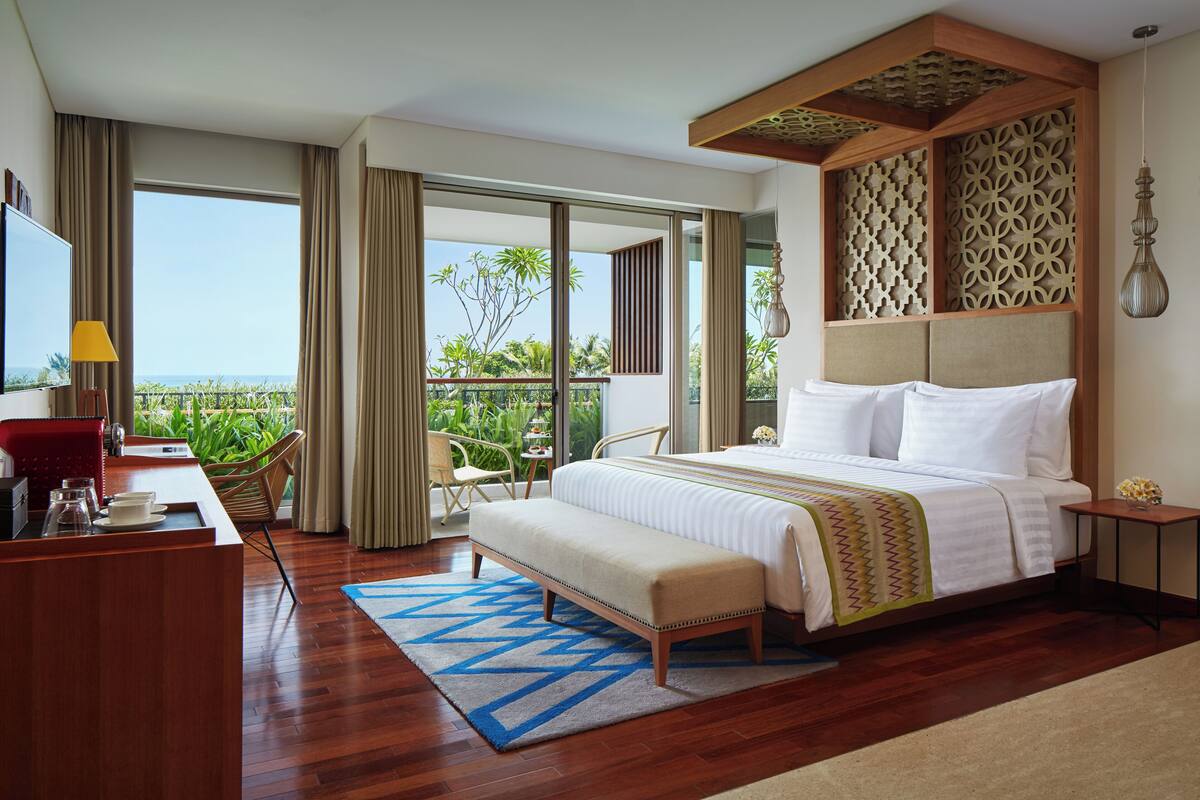 Habitación de hotel con una cama grande, un escritorio con lámpara, un banco y vistas a la vegetación a través de las puertas del balcón.