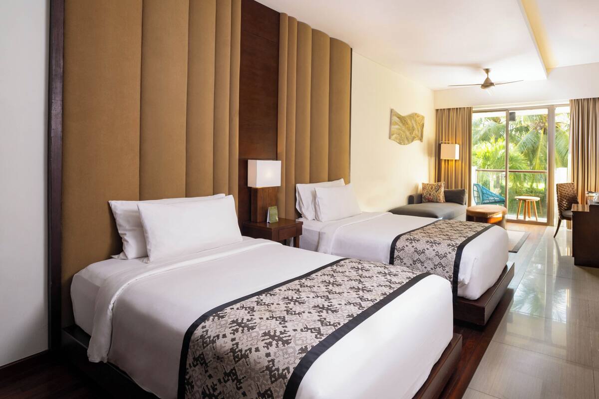 Habitación de hotel con dos camas, zona de estar, balcón con vistas y mostrador de recepción.