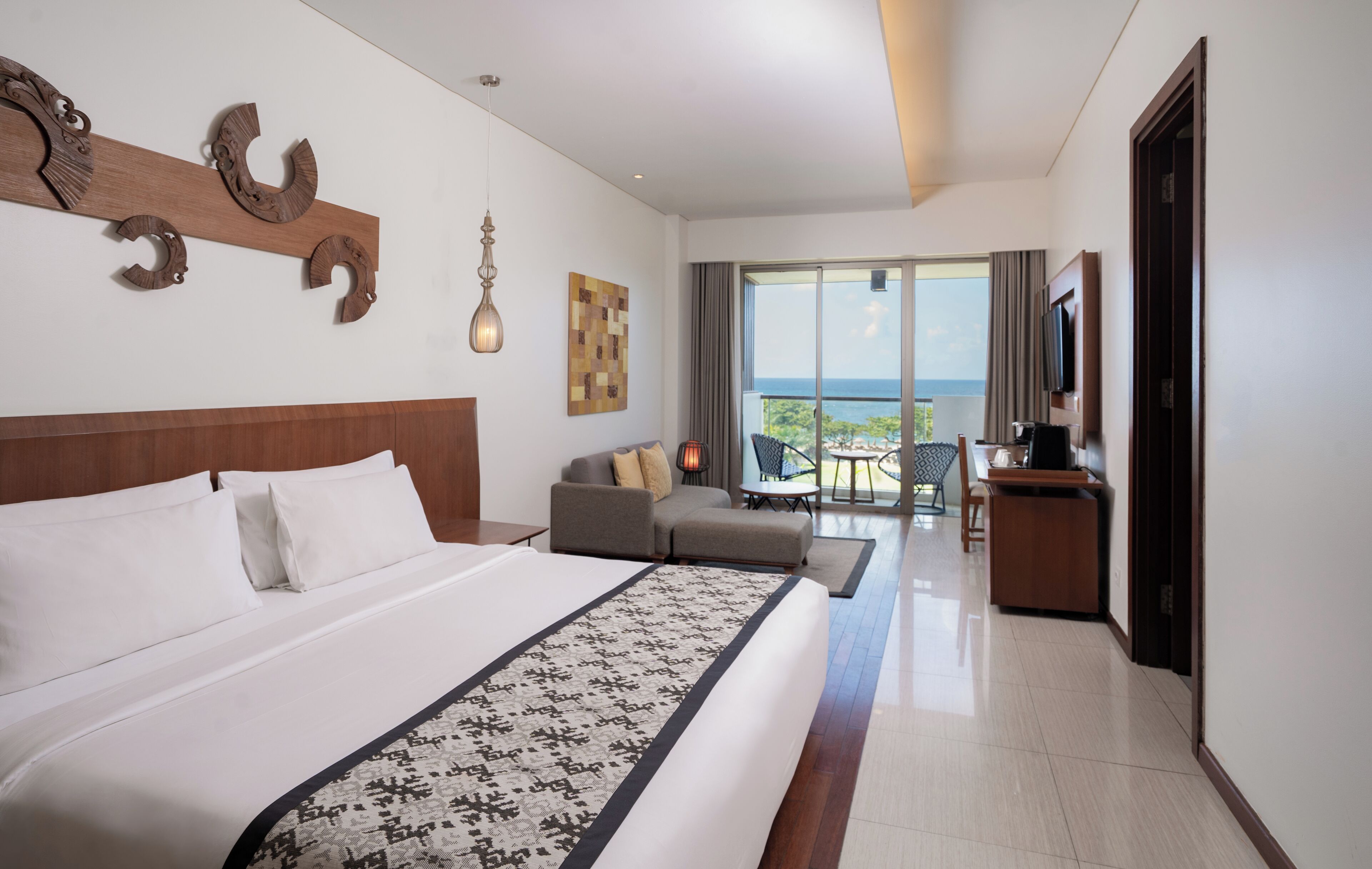 Una camera d'albergo con un letto grande, un divano, un tavolino e una TV. È presente un balcone con vista sul mare.
