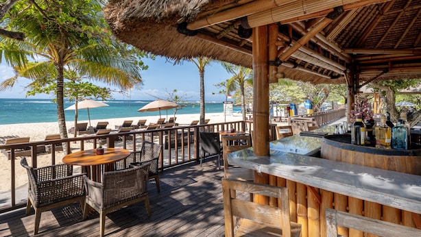 Una terraza de madera con mobiliario exterior, una zona de bar y vistas a la playa y el océano.