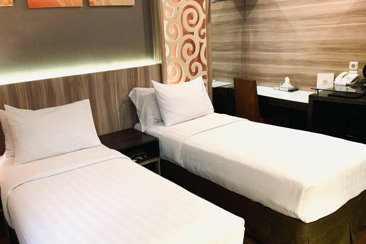 A hotel room with two single beds, a bedside table with a lamp, a decorative wall panel, and a desk with a telephone and a notepad.