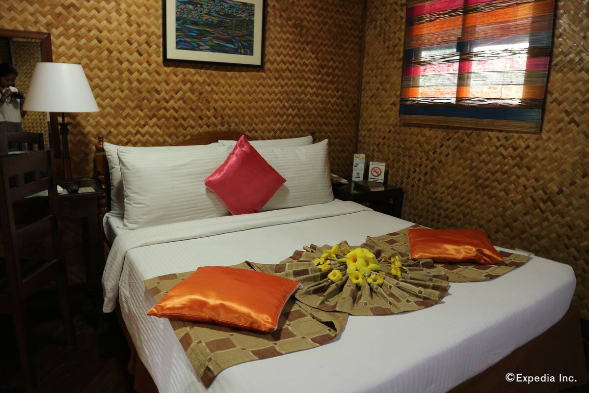 A hotel room with a bed, pillows, a lamp, a painting, and a window with blinds.