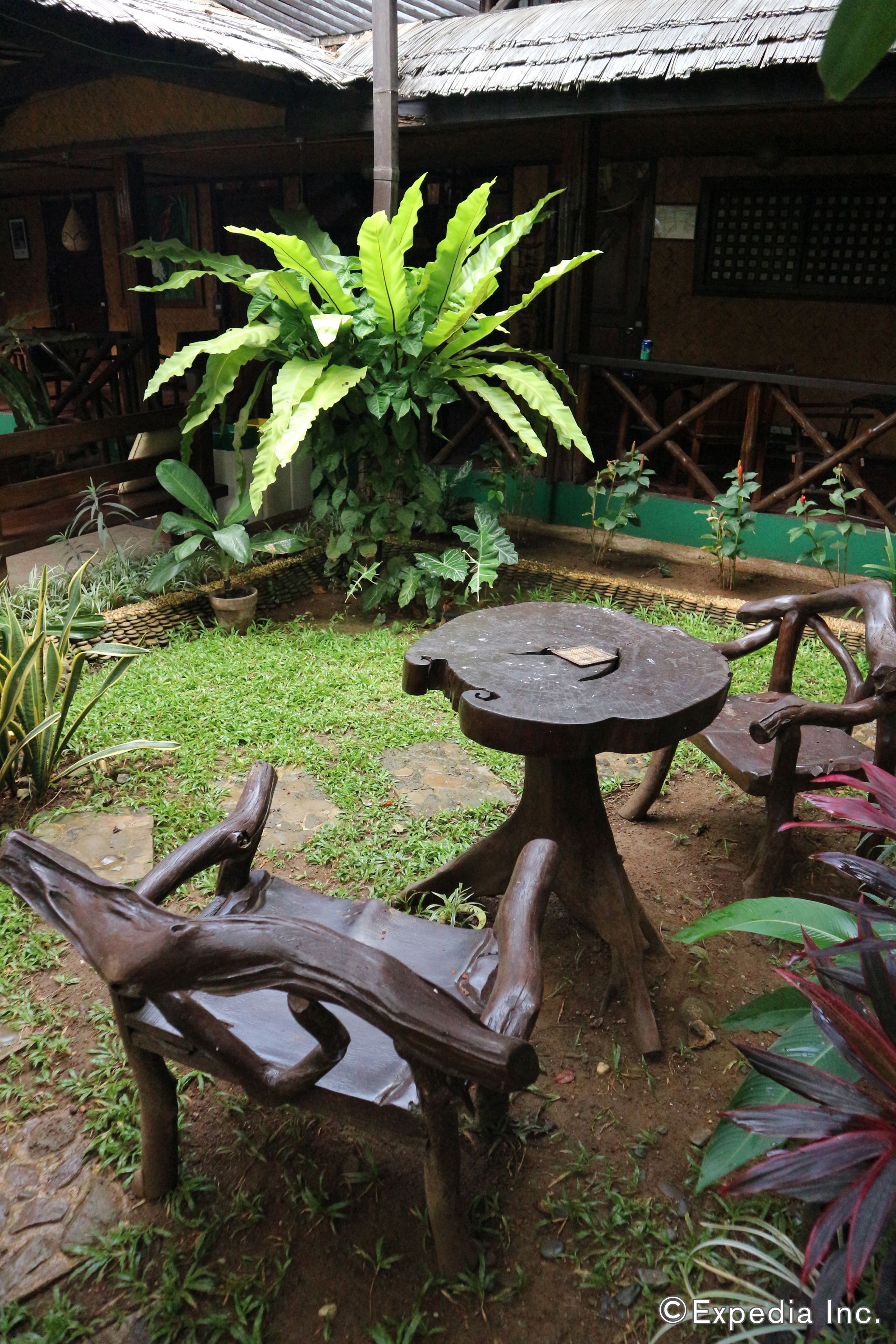 Un espacio de descanso al aire libre de estilo rústico, con una mesa de madera y bancos, rodeado de abundante vegetación y una estructura de techo de paja.