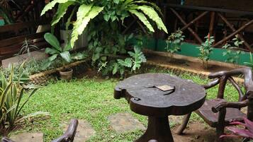 Un espacio de descanso al aire libre de estilo rústico, con una mesa de madera y bancos, rodeado de abundante vegetación y una estructura de techo de paja.