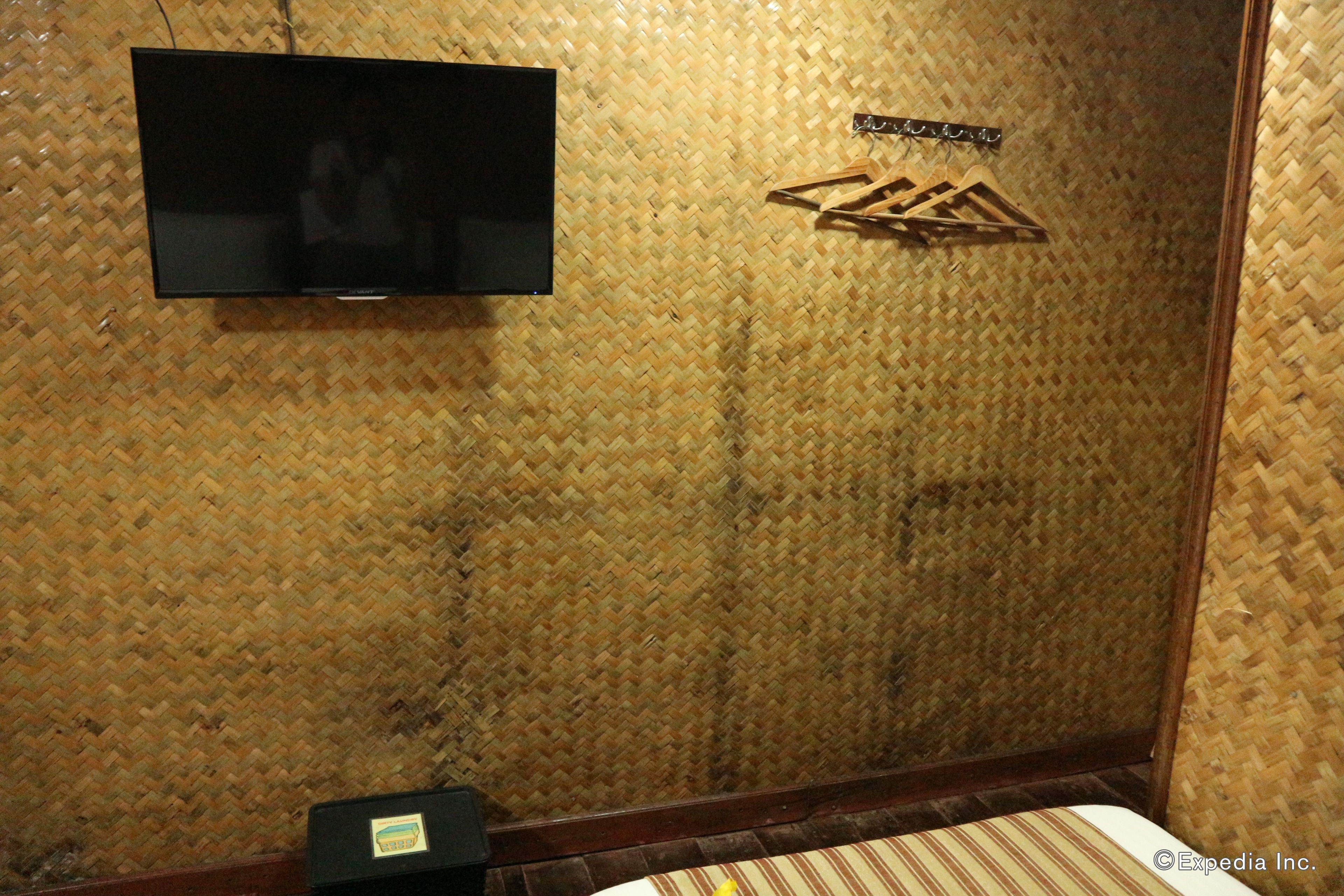 Une pièce avec un mur recouvert d’un revêtement tissé, un téléviseur à écran plat, un cintre en bois et un lit avec une literie à rayures.