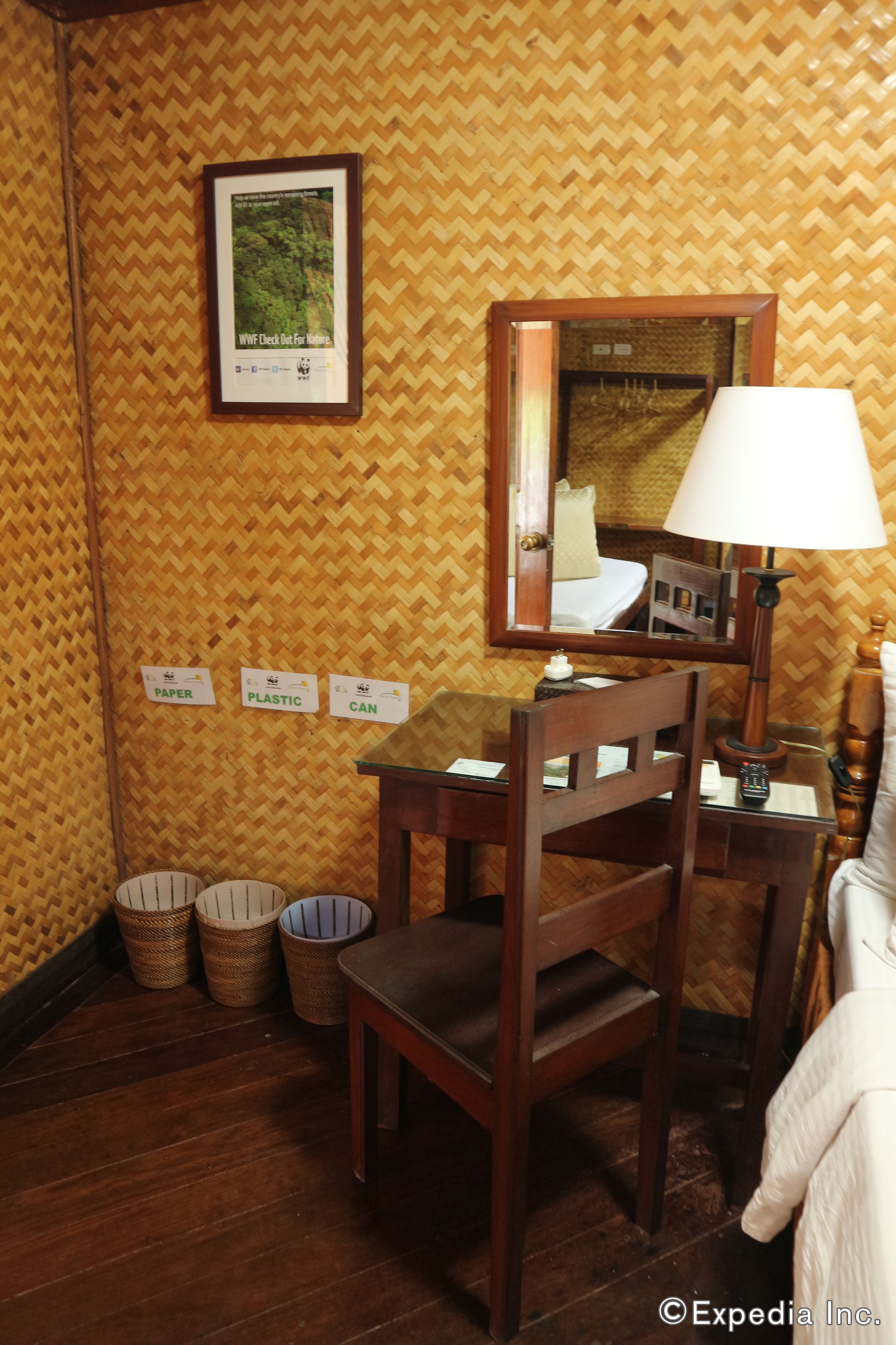 Habitación de hotel con un escritorio de madera, una silla, una lámpara y una cama. Se observan tres cestos de basura y un cuadro en la pared.