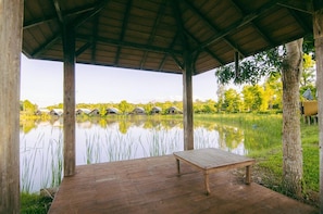 A wooden deck with a bench overlooking a calm lake, surrounded by trees and a thatched roof structure.
