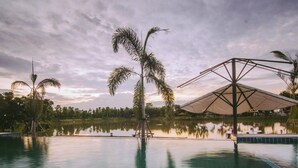 Un área de piscina tranquila con palmeras, un espacio de estar cubierto y una superficie de agua serena que refleja el entorno.