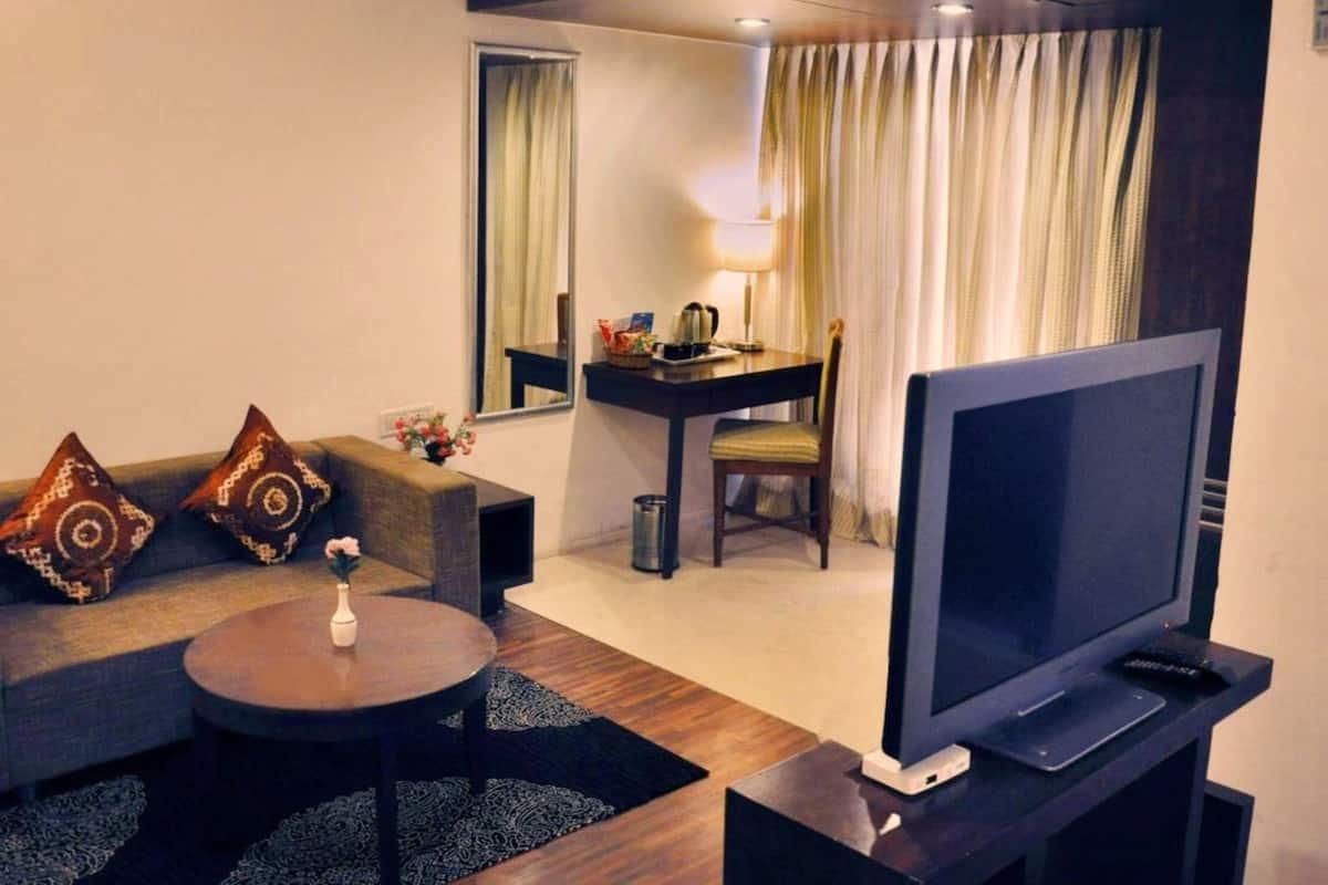 A modern hotel room with a flat-screen TV, a sofa with patterned cushions, a small round table, and a desk with a lamp.