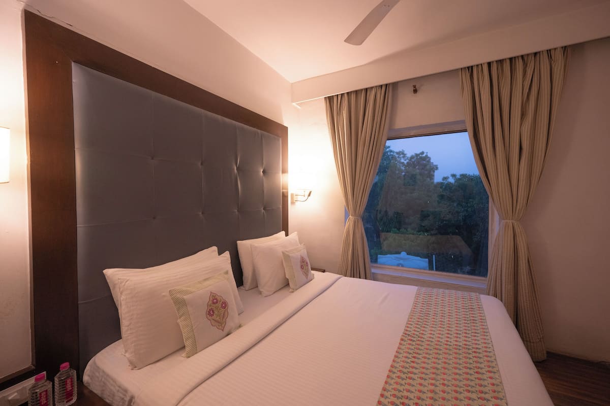 A hotel room with a large bed, a headboard, a window with curtains, and a nightstand with bottles.
