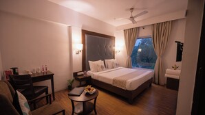 A hotel room with a bed, a desk, a chair, a small table with a laptop and fruit, and a window with curtains. - Hotel Clarks Collection Bhavnagar (Bhavnagar)