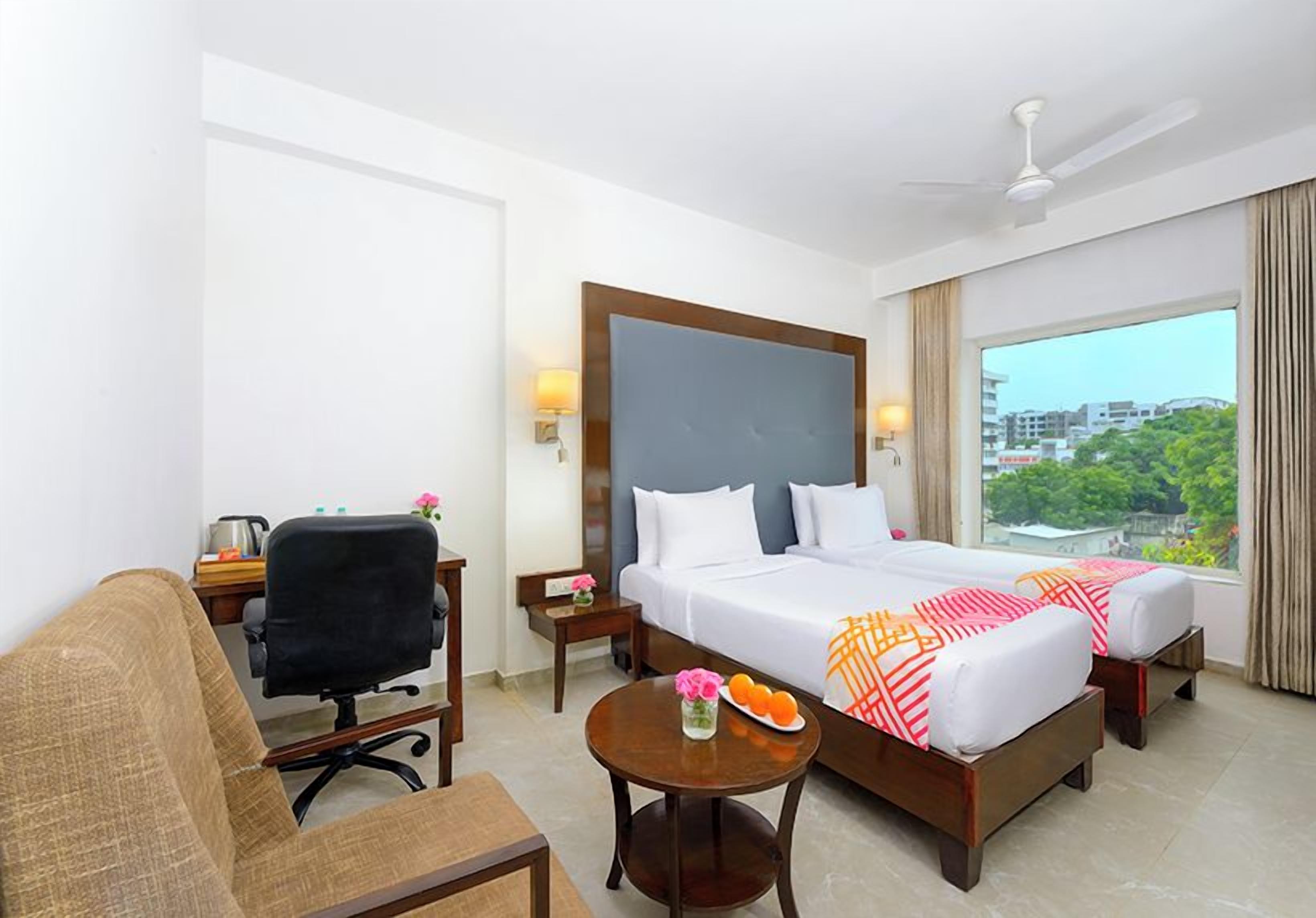 A hotel room with a bed, a desk, a chair, a small table with a vase and fruit, and a view of the city.