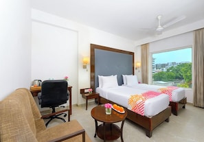 A hotel room with a bed, a desk, a chair, a small table with a vase and fruit, and a view of the city. - Ginger Bhavnagar, Kalubha Road (Bhavnagar)