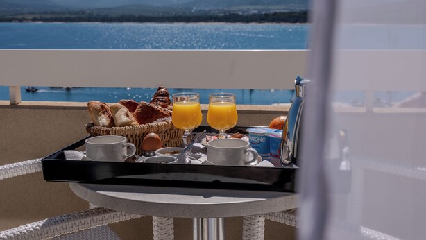 A breakfast tray with bread, eggs, juice, and a box of cereal on a balcony overlooking a body of water and mountains.