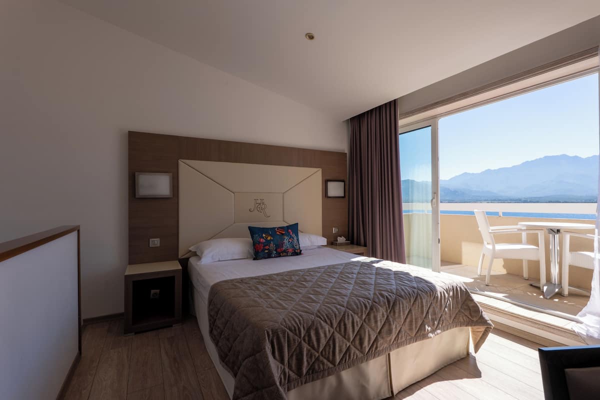Una habitación de hotel con cama, mesita de noche, ventana con vista a las montañas y balcón con mesa y sillas.