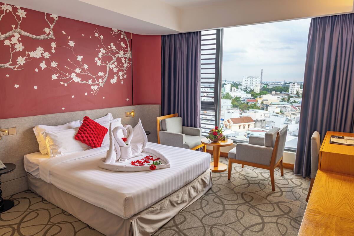 A hotel room with a large bed, a desk, a chair, and a view of the city through large windows.