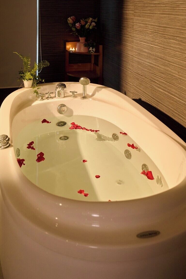 A bathtub with rose petals floating on the water, a small potted plant, and a candle on a table in the background.