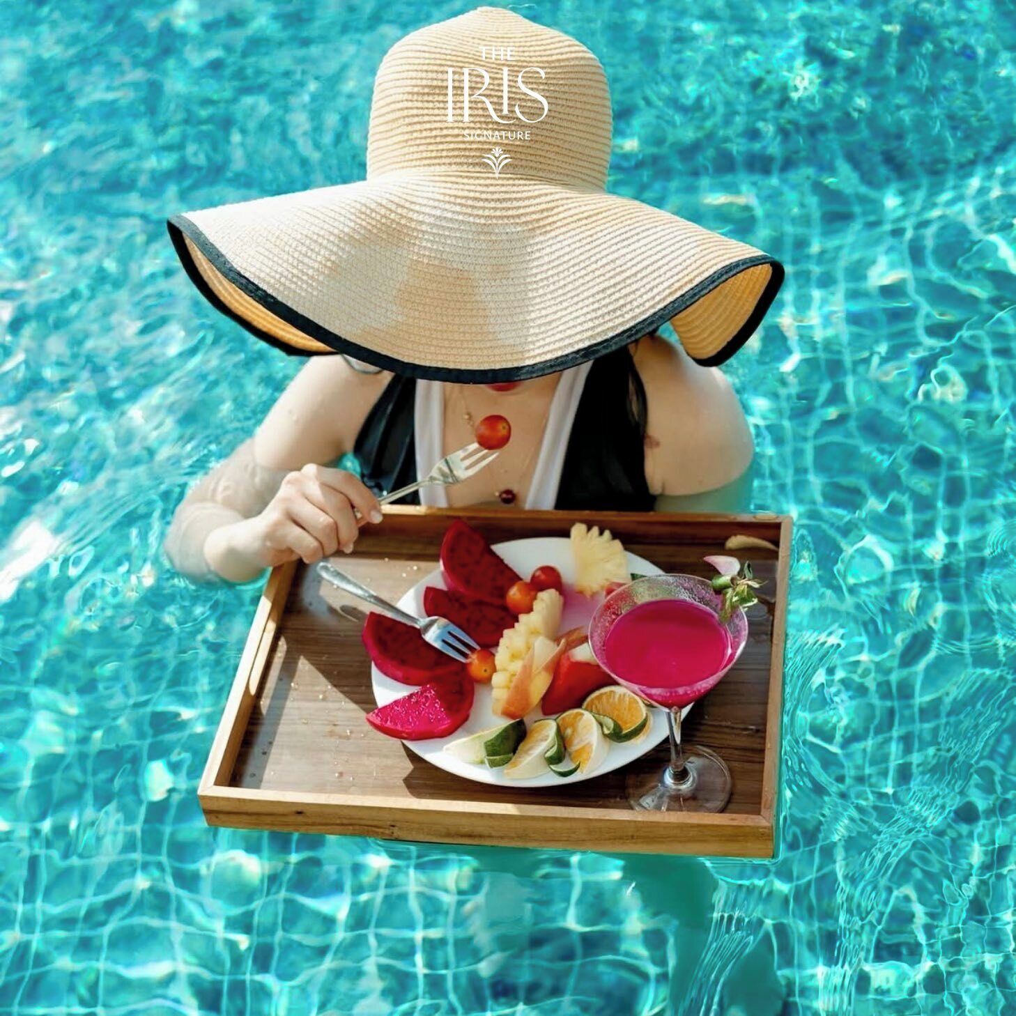 Una persona en una piscina, usando un sombrero de paja, sosteniendo una bandeja con comida y una bebida.