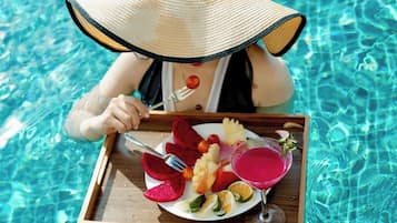 Una persona en una piscina, usando un sombrero de paja, sosteniendo una bandeja con comida y una bebida.