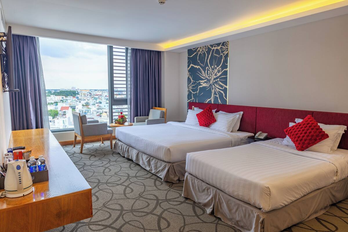 A hotel room with two beds, a desk with a coffee maker, a chair, and a view of the city.