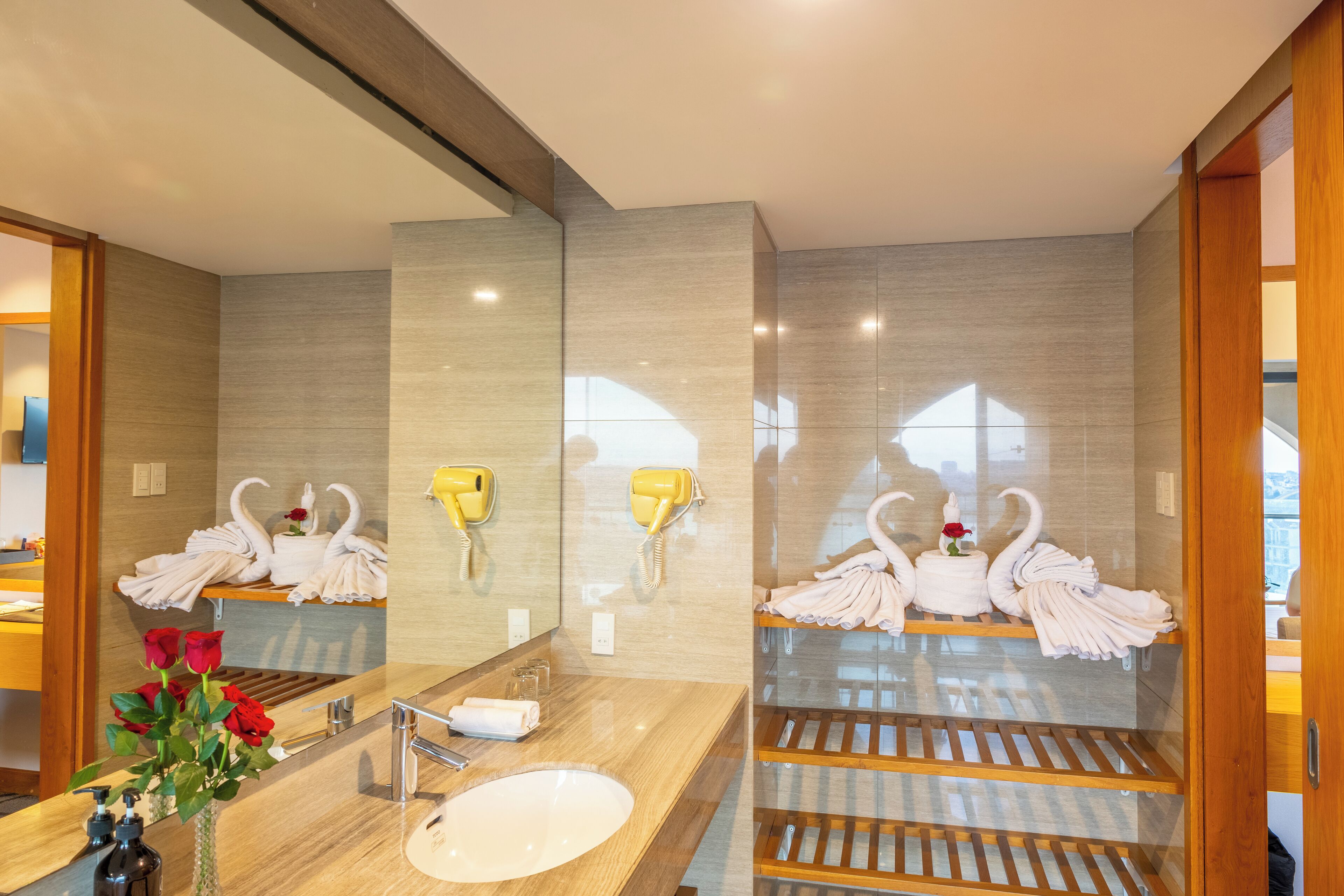Une salle de bain moderne avec un grand miroir, deux sèche-cheveux fixés au mur, un meuble-lavabo en bois et une cabine de douche en verre.