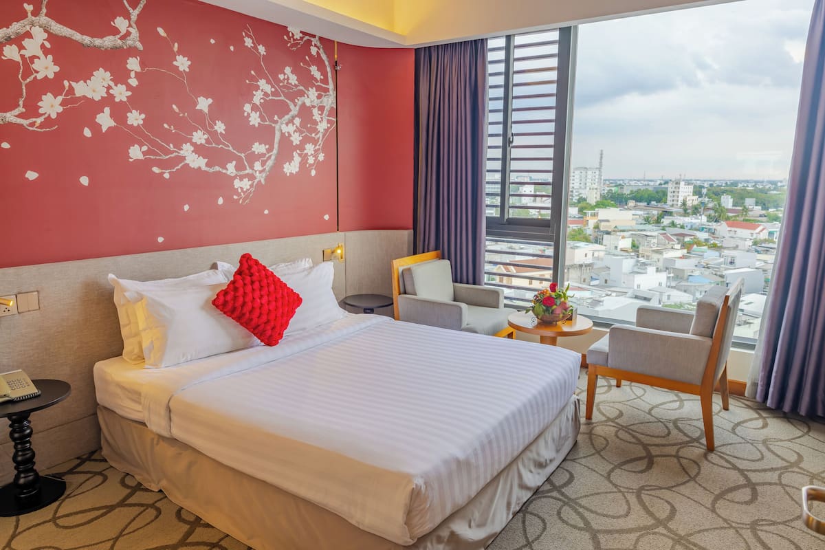 A hotel room with a large bed, a desk, a chair, and a view of the city through the window.