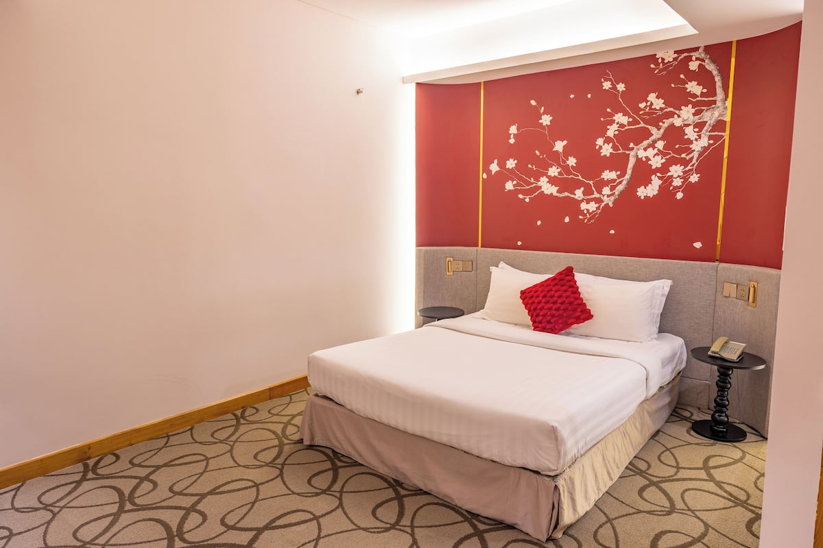 A hotel room with a bed, a red decorative wall, a patterned carpet, and a bedside table with a phone.