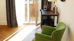 Una habitación con piso de madera, un sillón verde, una mesa con lámpara y un televisor en la pared.