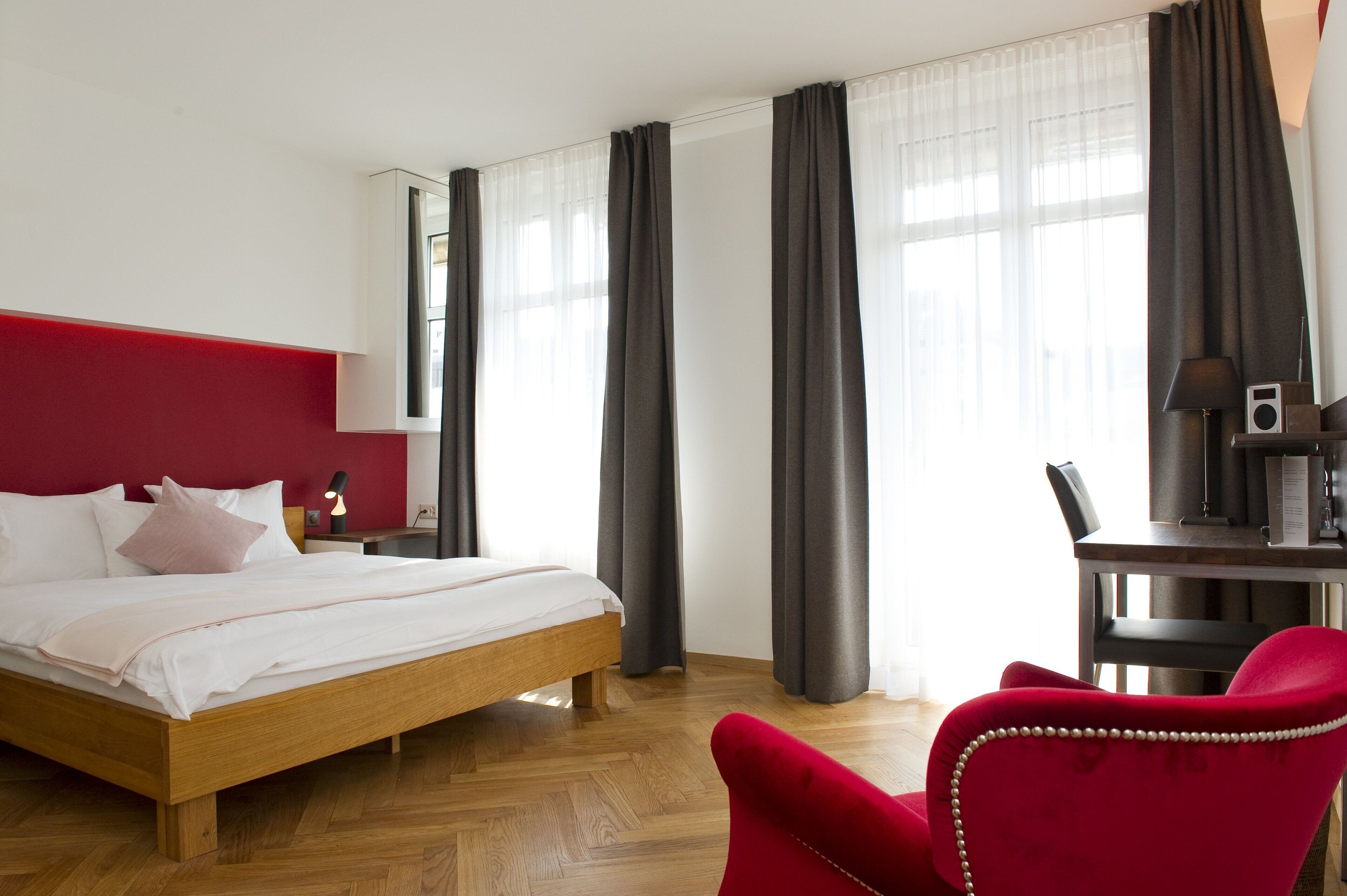 Una habitación de hotel con una cama de madera, una silla roja, un escritorio con lámpara y un ventanal con cortinas.