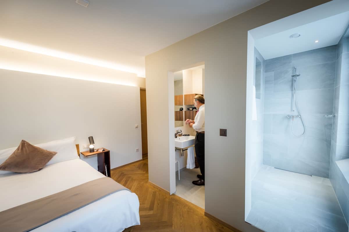 A modern hotel room with a bed, a nightstand, a bathroom with a shower, and a visible kitchen area.