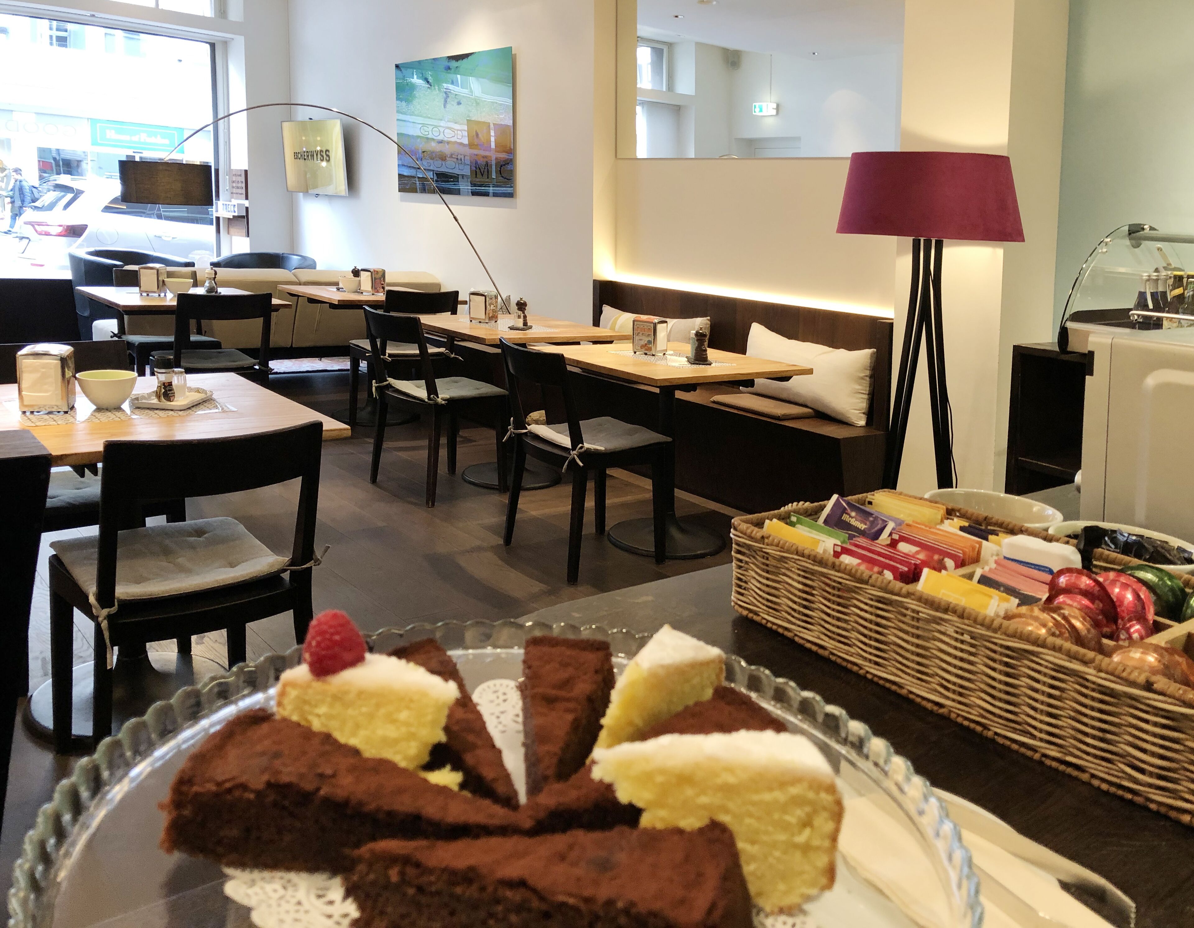 Een gezellig café met houten tafels en stoelen, een rieten mand met lekkernijen en een glazen vitrine met brownies.