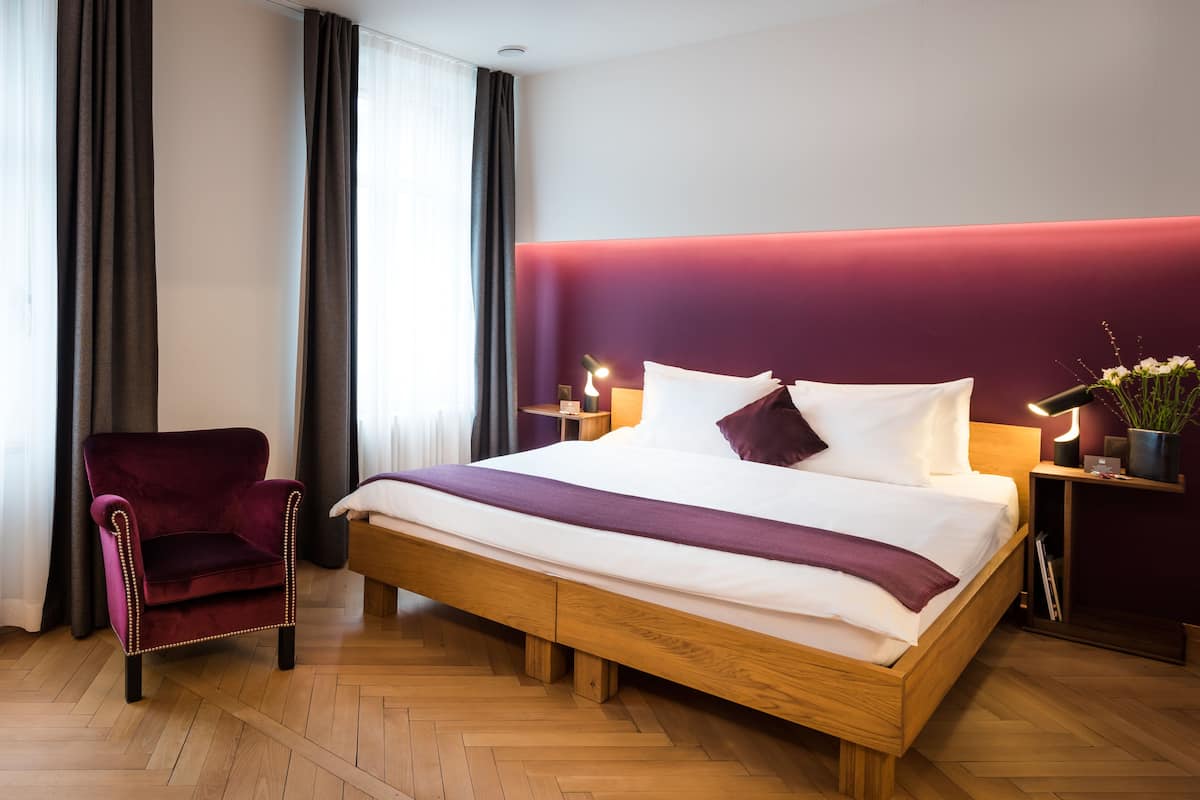 A modern hotel room with a wooden bed, a red armchair, and a nightstand with a lamp.
