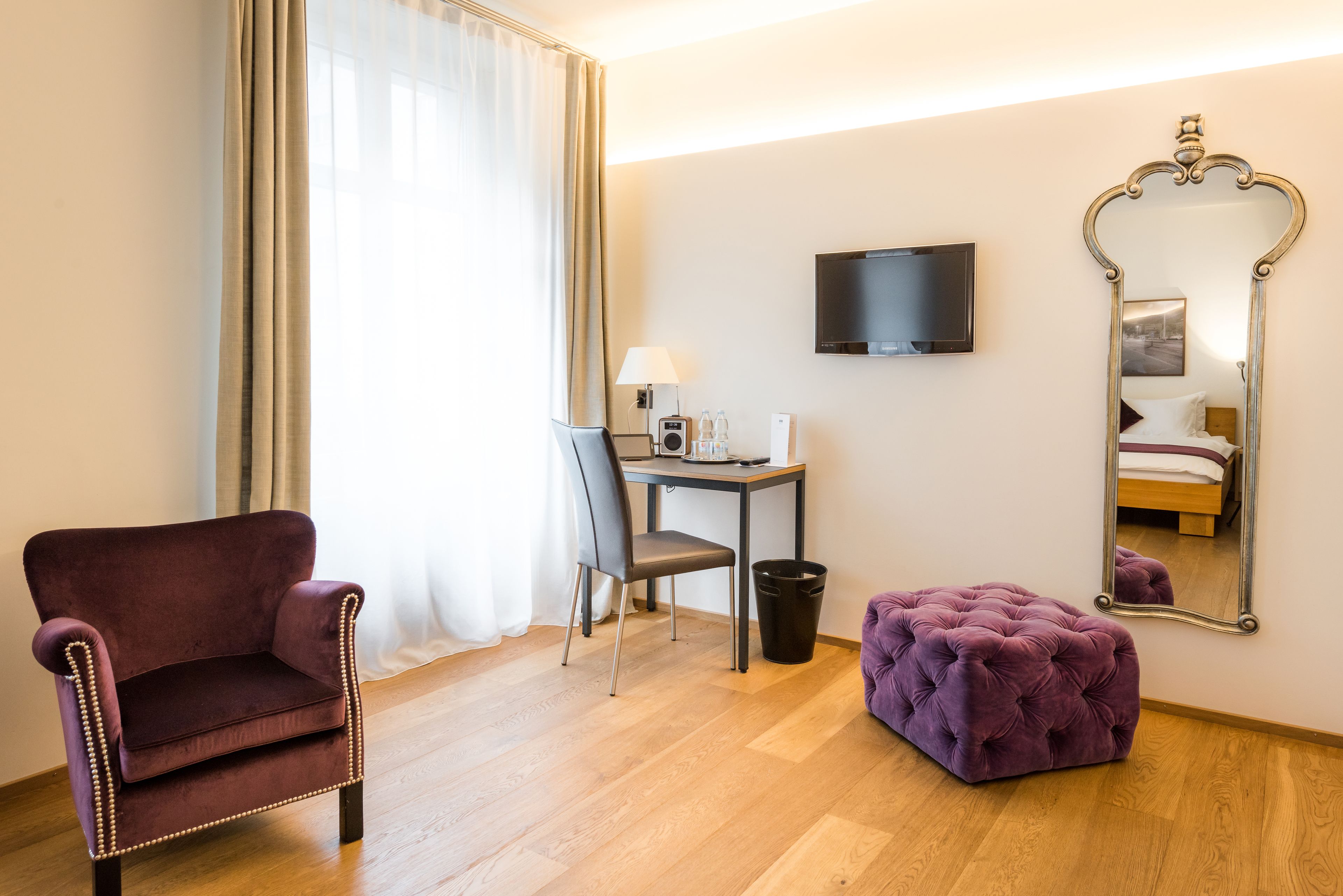 Ein Hotelzimmer mit Holzboden, einem dunkelroten Sessel, einem kleinen Tisch mit Lampe, einem Fernseher an der Wand und einem großen Spiegel.