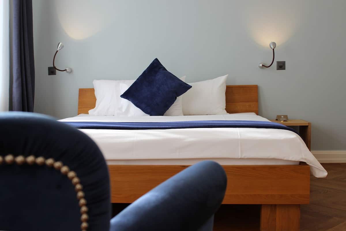 A neatly made bed with white linens and a blue accent pillow, a wooden headboard, and a bedside table with a lamp.