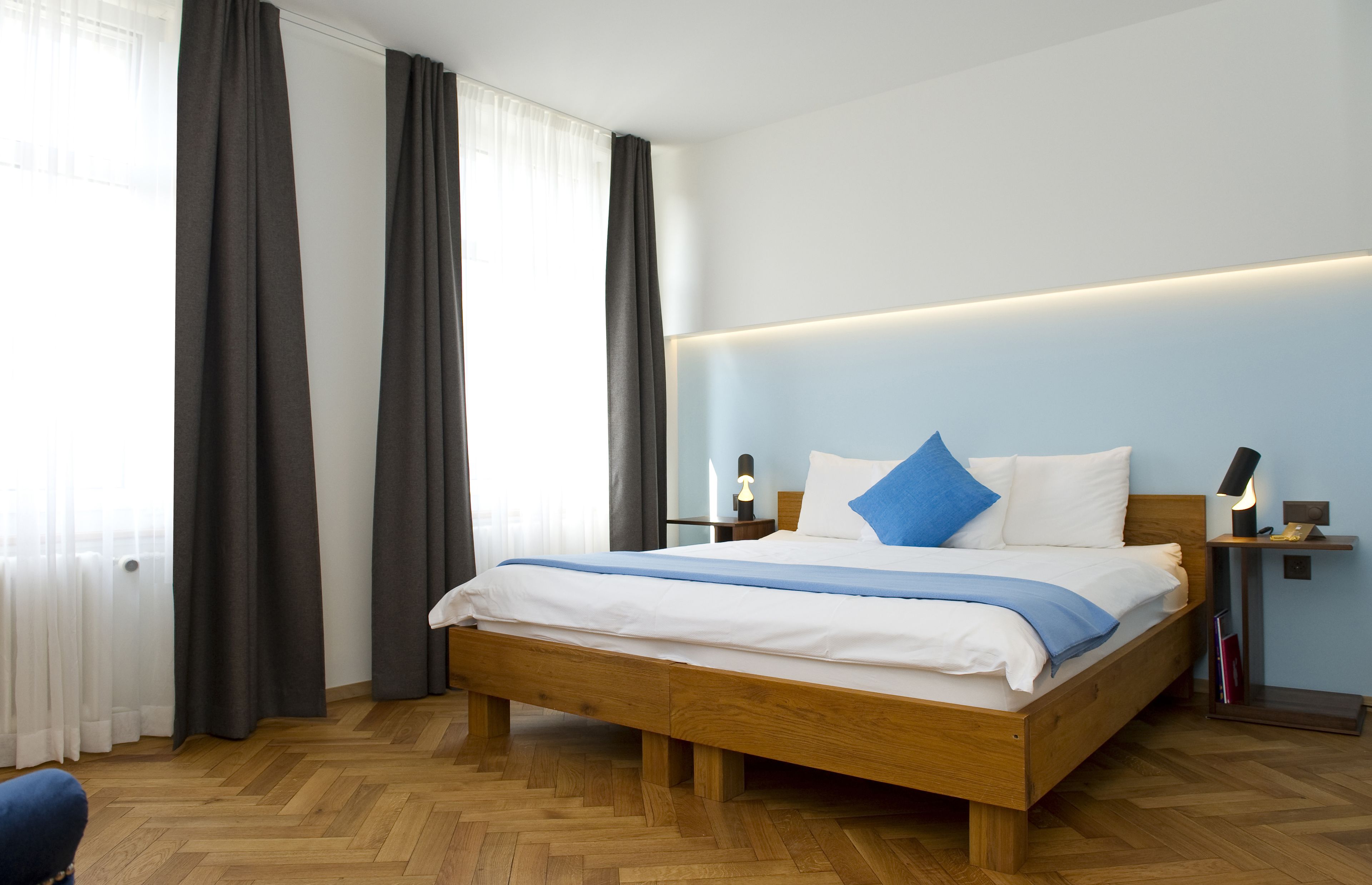 A modern hotel room with a wooden bed, a nightstand with a lamp, and a view of the city through large windows.