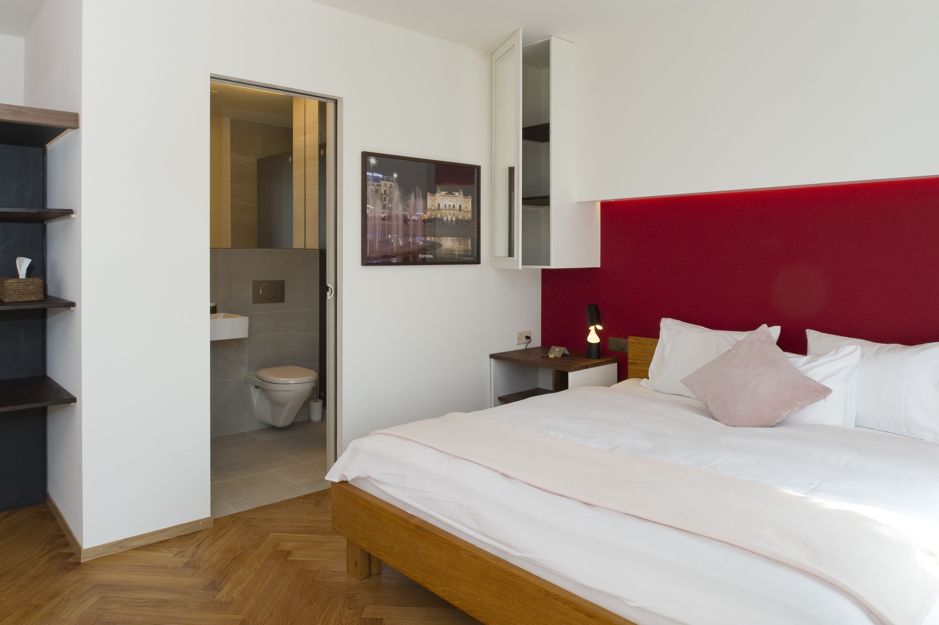 Une chambre d’hôtel moderne avec un lit en bois, du linge de lit blanc, un mur d’accent rouge, un tableau encadré et une petite table de chevet avec une lampe.