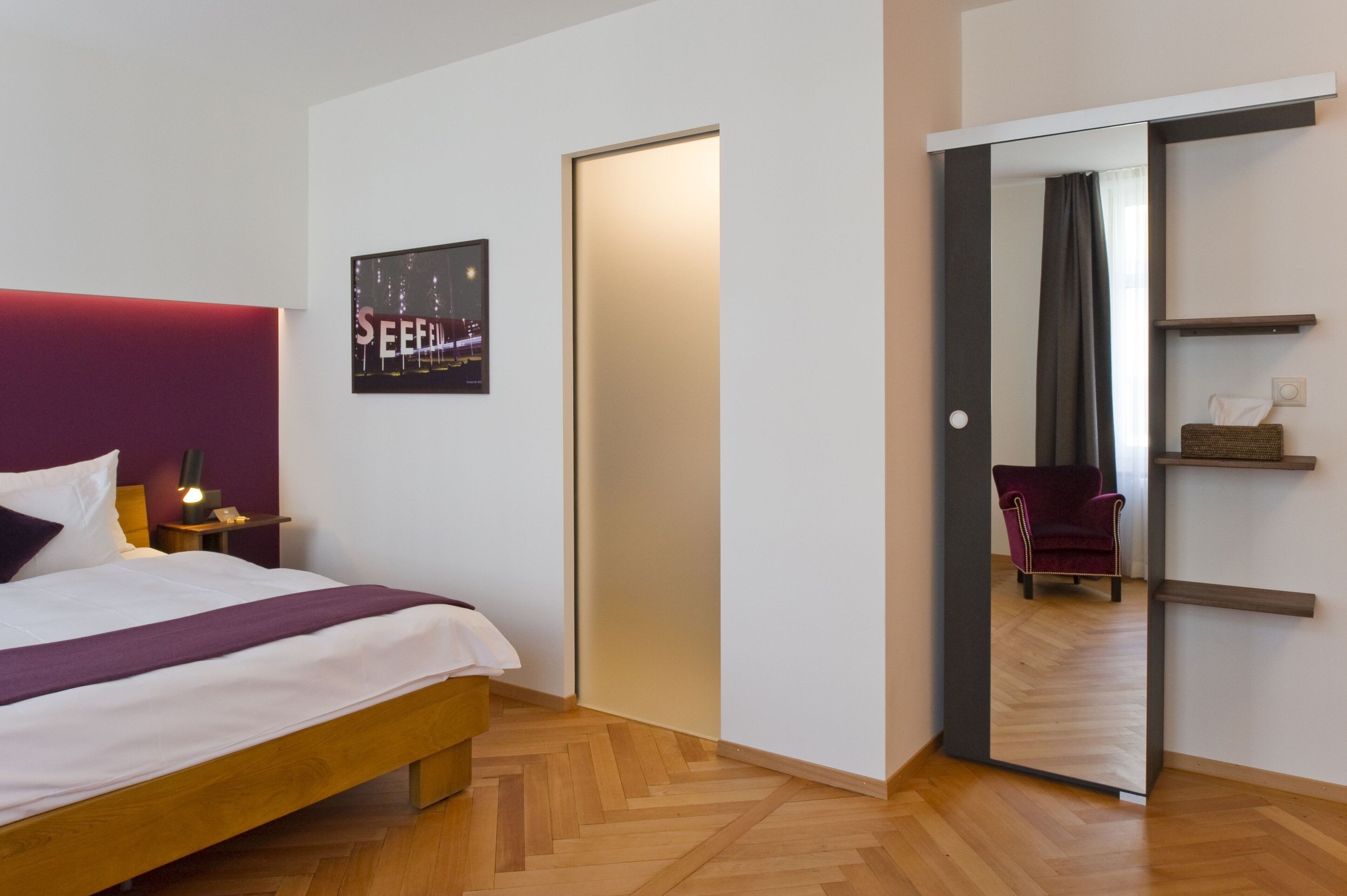 Een hotelkamer met een bed, nachtkastje, spiegel, deur en stoel.