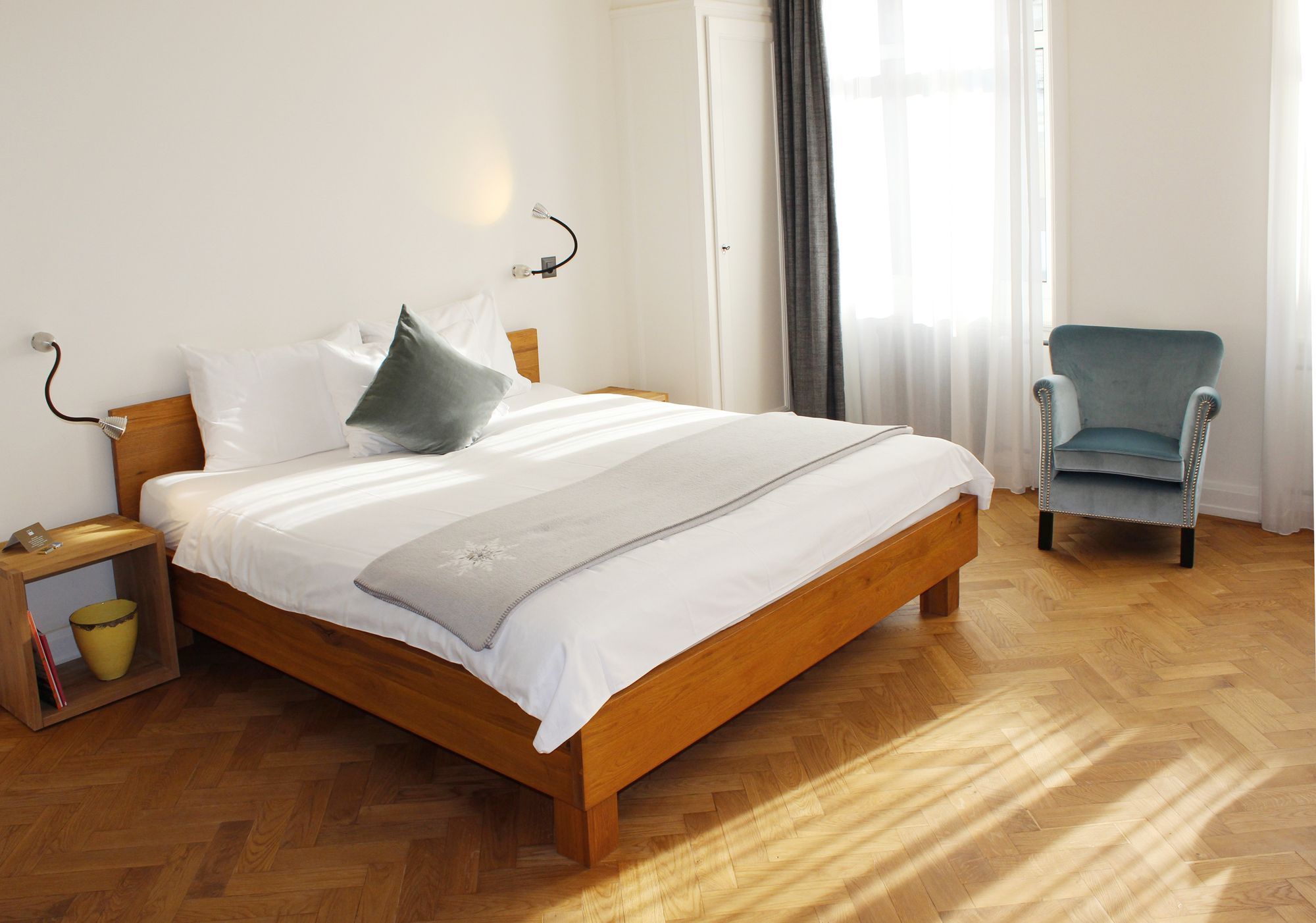A neatly made bed with a wooden frame, a bedside table with a lamp, and a chair in a bright room with wooden flooring.