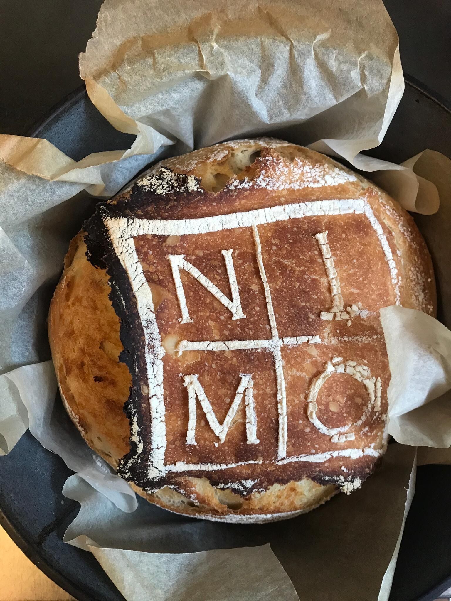 A round bread with a grid pattern and letters on top, placed in a black container with parchment paper.