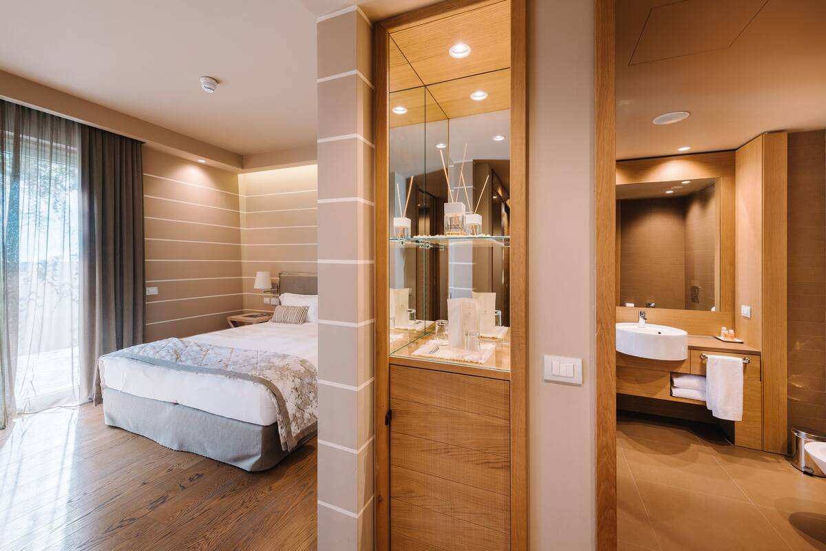 Une chambre d’hôtel moderne dotée d’un grand lit, d’une coiffeuse en verre et d’une salle de bain séparée avec un lavabo et un miroir.