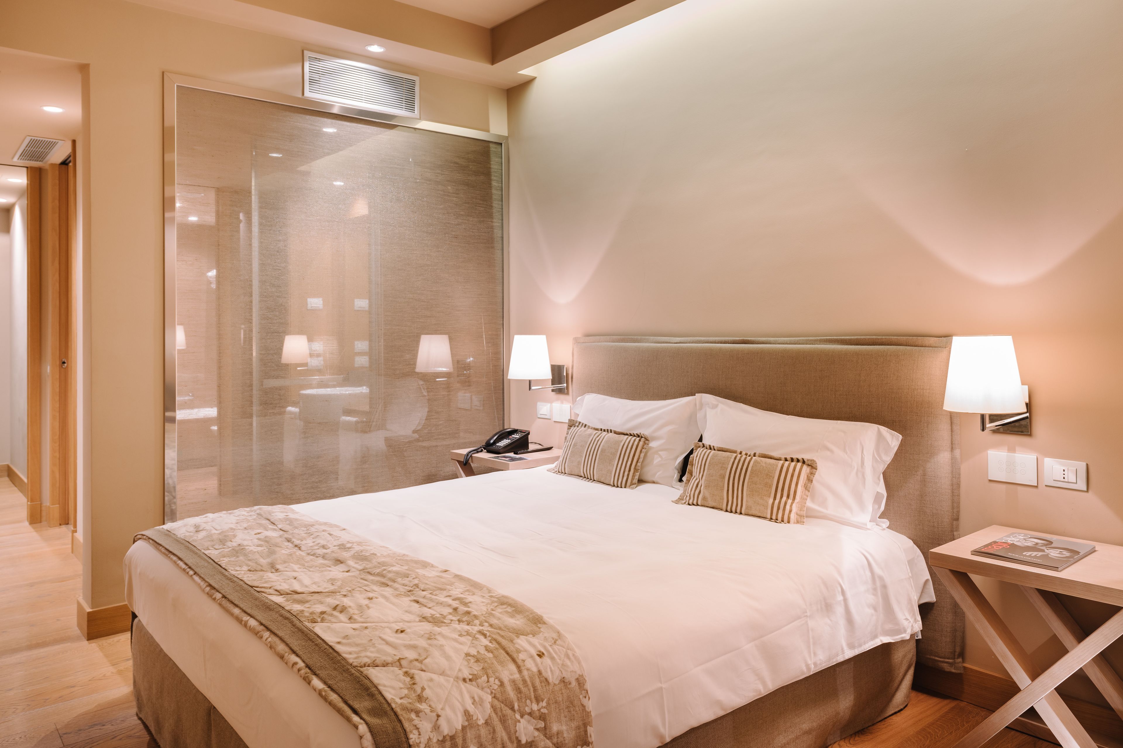 A hotel room with a large bed, bedside lamps, a nightstand, and a glass partition.