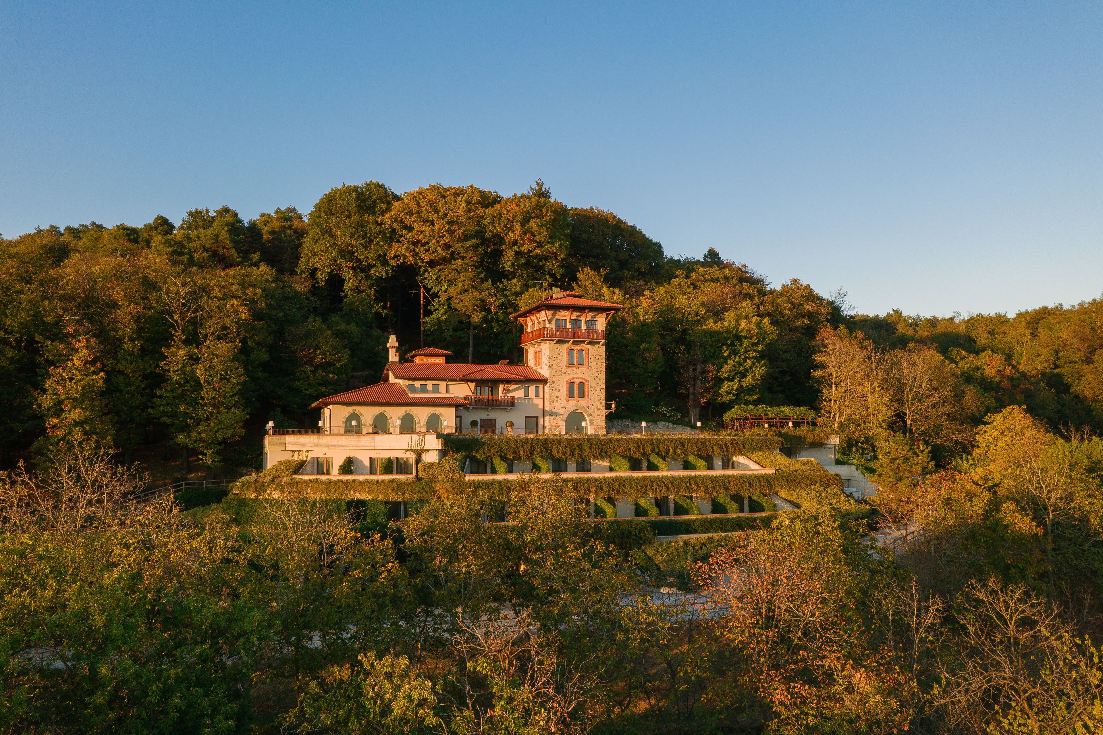 A large, historic building with a tower, surrounded by dense forest and a well-maintained garden.