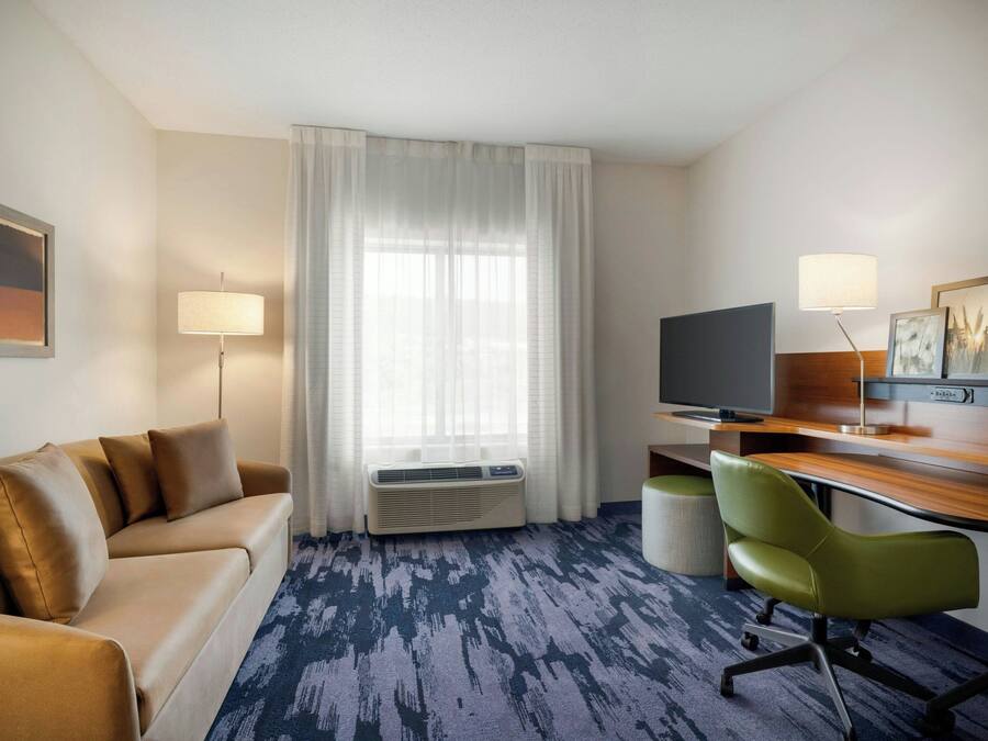A hotel room with a sofa, a desk with a computer, a chair, a TV, and a large window with curtains.