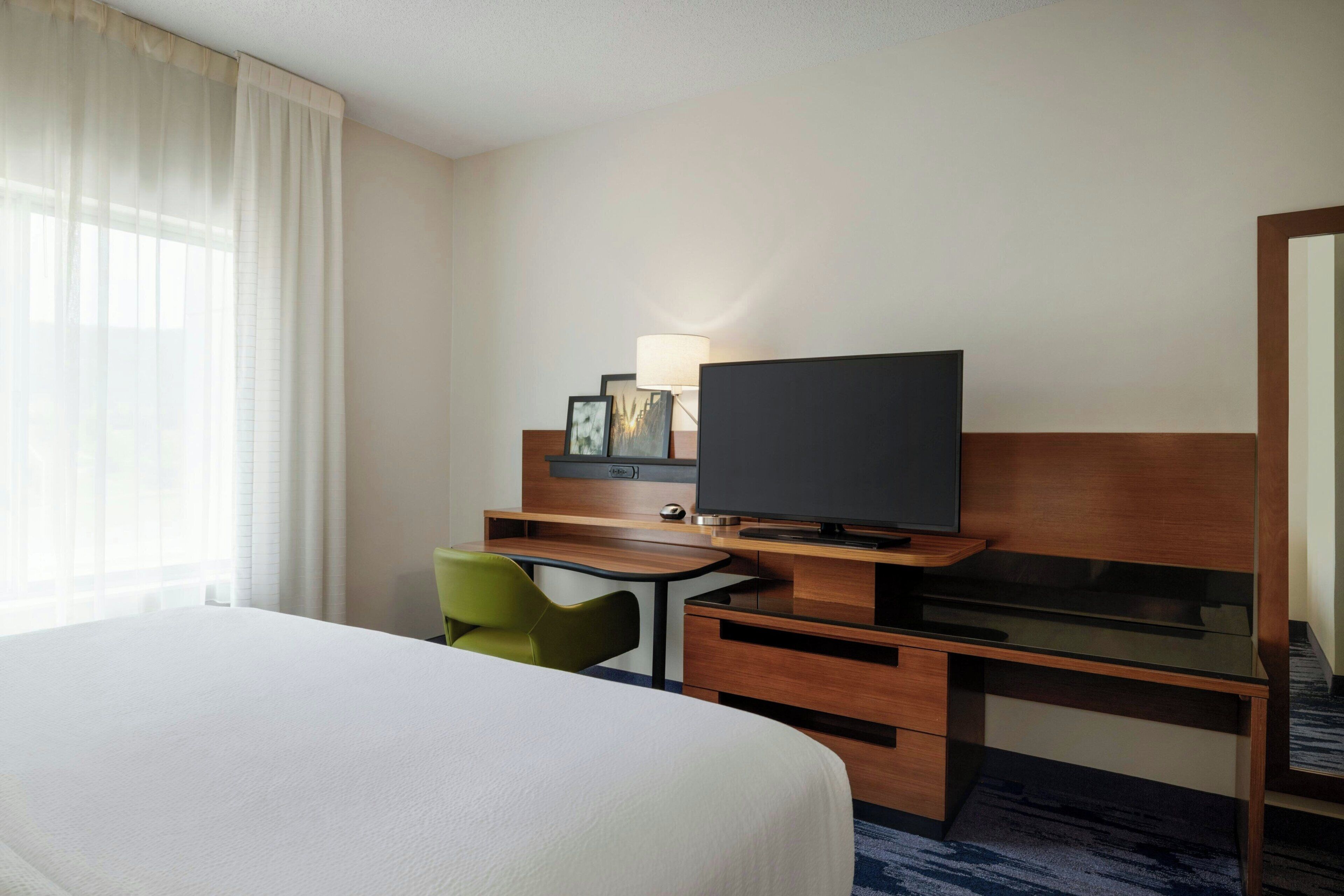 A hotel room with a large bed, a desk with a flat-screen TV, a chair, and a lamp.