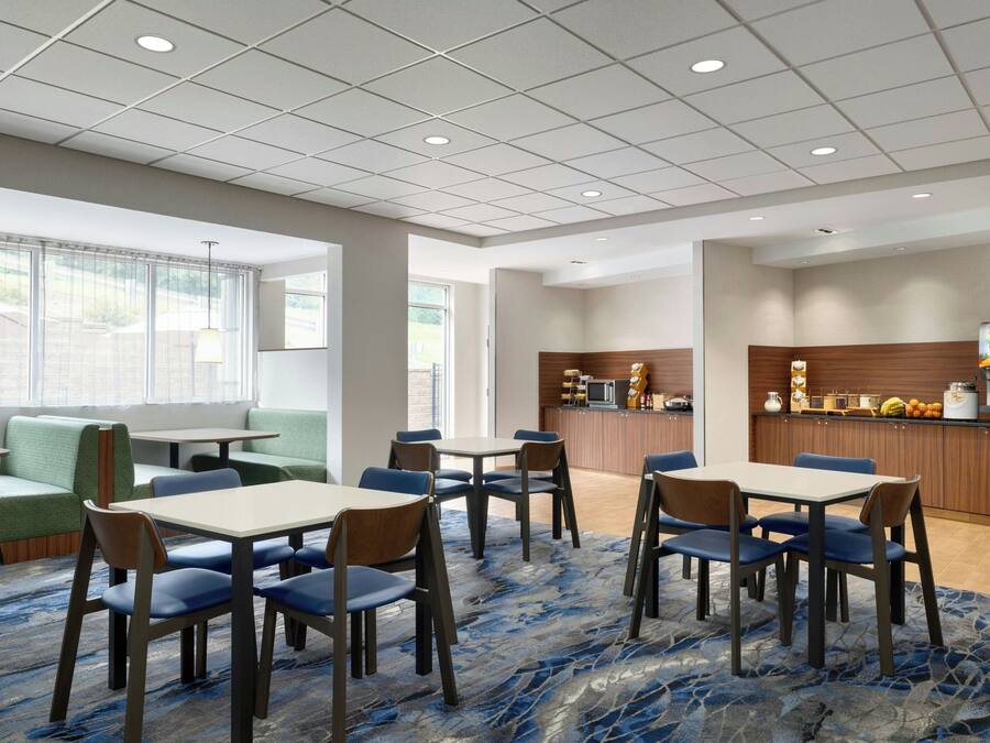 A hotel dining area with tables and chairs, a kitchen area, and a seating area with a view of the outdoors.