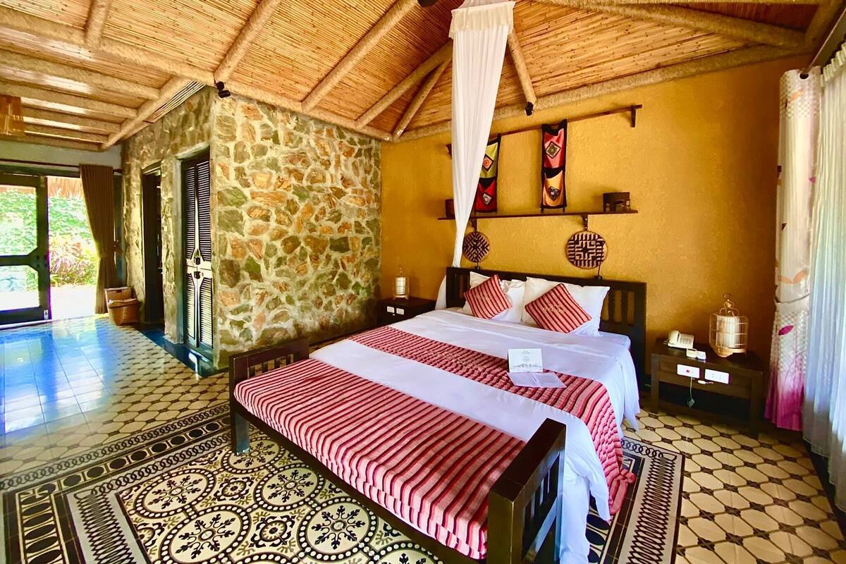 Een kamer met een bed, stenen muren, een rieten dak en een kamer met een versierd vloerkleed.