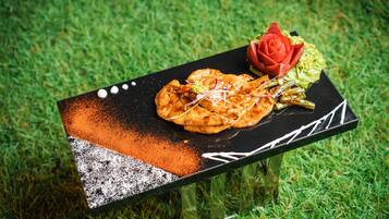 Een zwart rechthoekig tafelkleed met een decoratief patroon, een bord met eten, een rode roos en wat groen op een gazon.