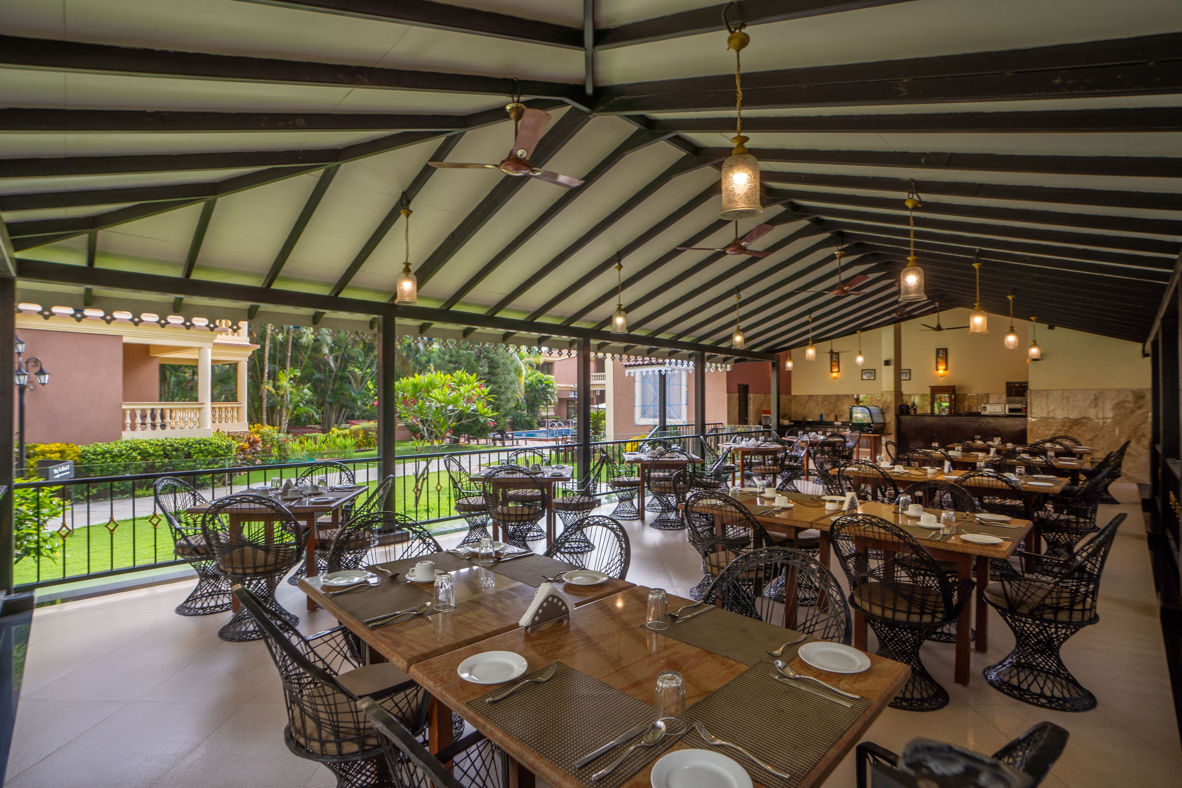 An outdoor dining area with tables and chairs, a covered roof, and a view of the garden.